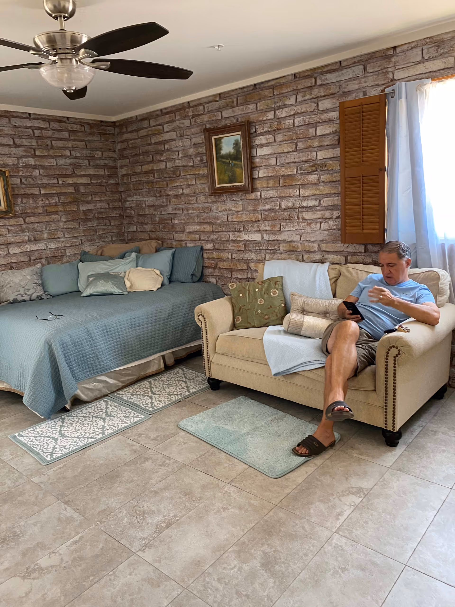 A man sitting on a beige couch in a room with brick walls and tiled floor. The room has a ceiling fan, a bed with blue bedding and multiple pillows, two framed paintings on the wall, and a window with white curtains and wooden shutters.