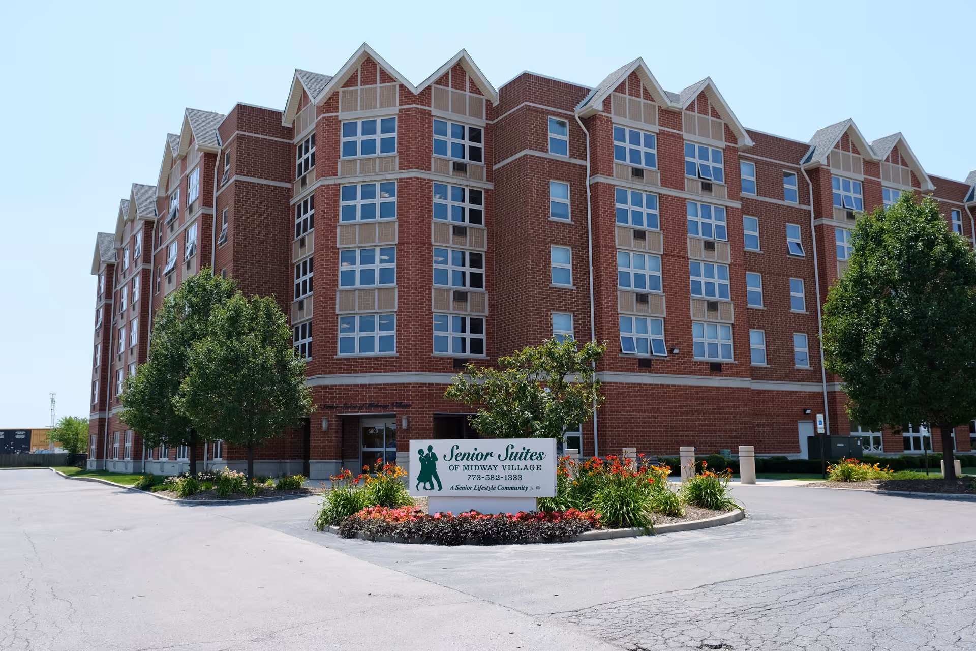 Exterior view of a large brick senior living facility building with multiple windows and peaked roof sections. In front of the building is a landscaped roundabout with flowers and greenery, featuring a sign that reads 'Senior Suites of Midway Village' along with a phone number and the phrase 'A Senior Lifestyle Community.'