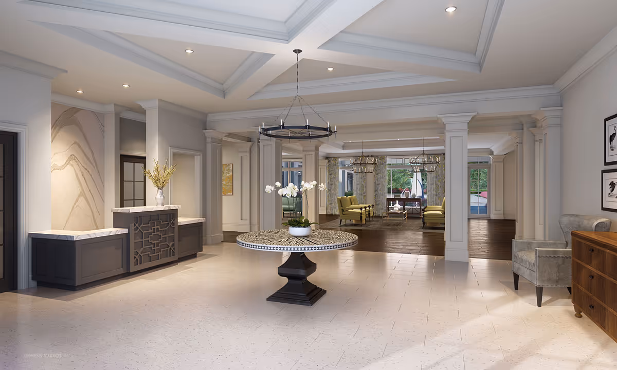 Spacious and elegant interior common area with a round table featuring a floral centerpiece in the center. The room has a high tray ceiling with recessed lighting and a chandelier. To the left, there is a built-in cabinet with a vase of flowers. In the background, there is a seating area with green armchairs and large windows letting in natural light. The floor transitions from light tile in the foreground to dark wood in the seating area.