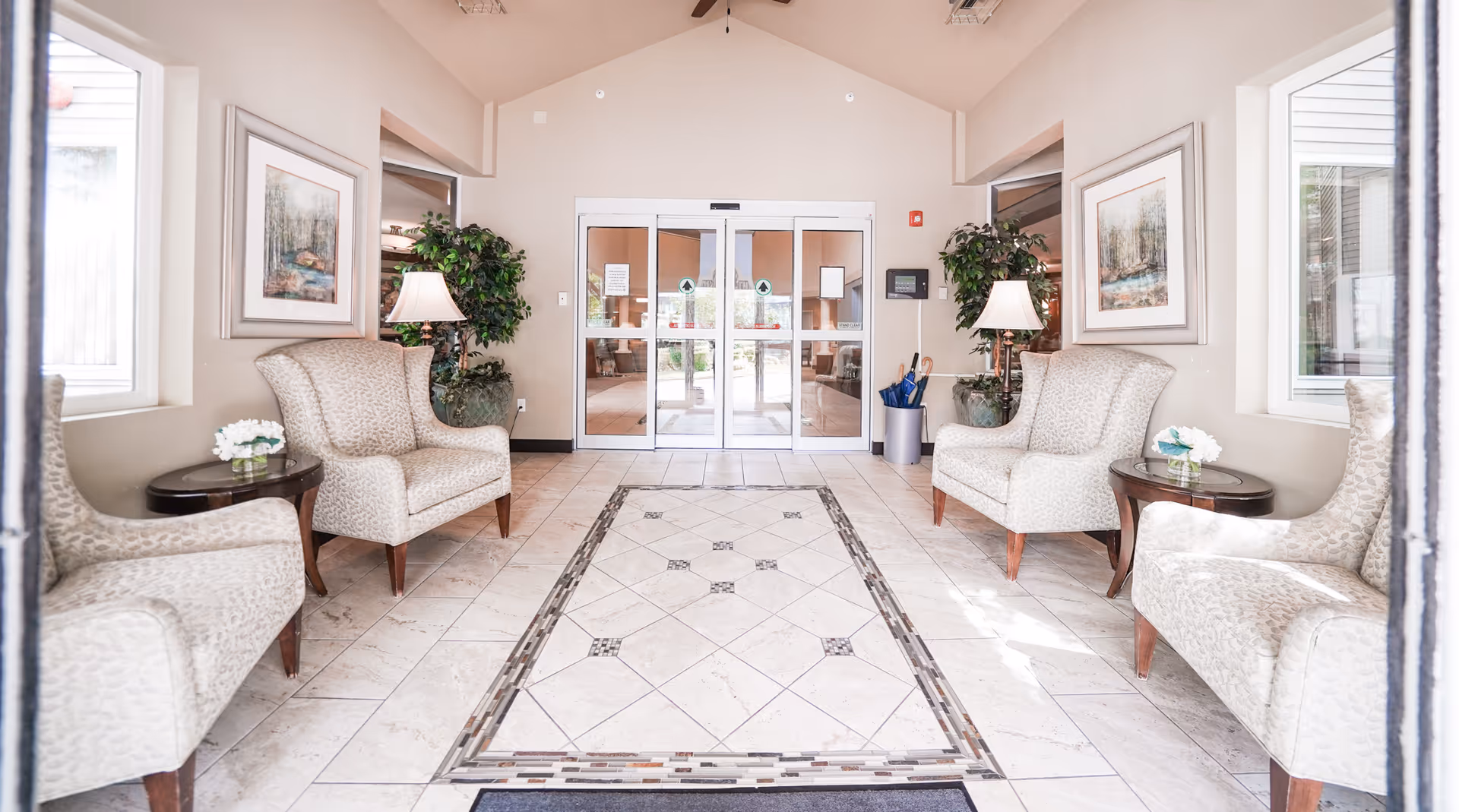Bright and welcoming lobby area with four patterned armchairs arranged around two small round tables with white flower arrangements. Two table lamps and two large potted plants flank the double glass entrance doors. The floor features a decorative tile design and the walls are adorned with framed artwork.