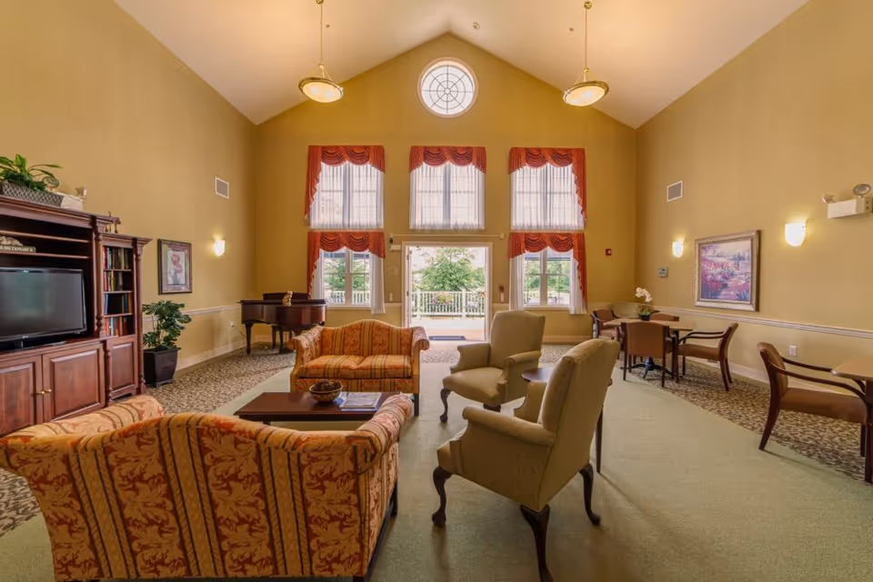 A spacious and well-lit living room in an assisted living facility featuring high vaulted ceilings, large windows with red valances, and a glass door leading outside. The room is furnished with patterned sofas, armchairs, a wooden coffee table, a grand piano, a TV on a wooden entertainment center, and several small tables with chairs. The walls are painted a warm beige color and decorated with framed artwork and plants.
