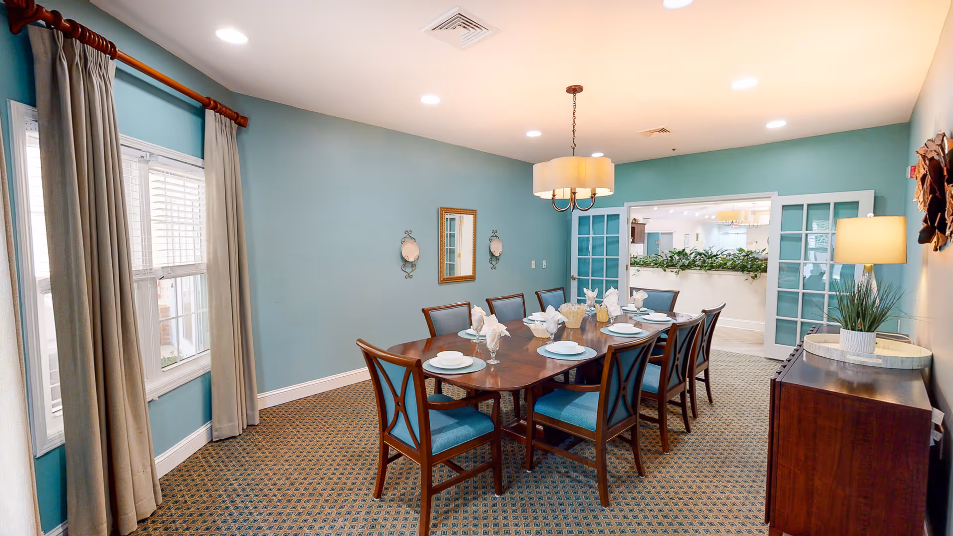 A dining room with a long wooden table set for eight people with plates, glasses, and napkins. The room has teal walls, a patterned carpet, beige curtains on a window, and a chandelier hanging above the table. There is a wooden sideboard with a lamp and a plant on the right side, and double glass doors leading to another room.