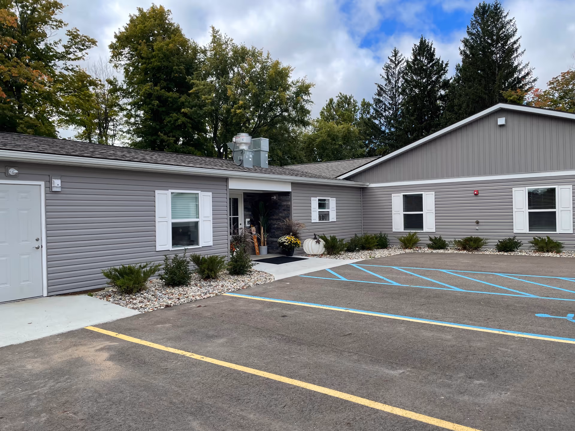 Exterior view of a single-story building with gray siding and white trim, surrounded by small bushes and decorative rocks. There is a parking lot with marked spaces, including a handicapped spot, in front of the building. Trees with green foliage are visible in the background under a partly cloudy sky.