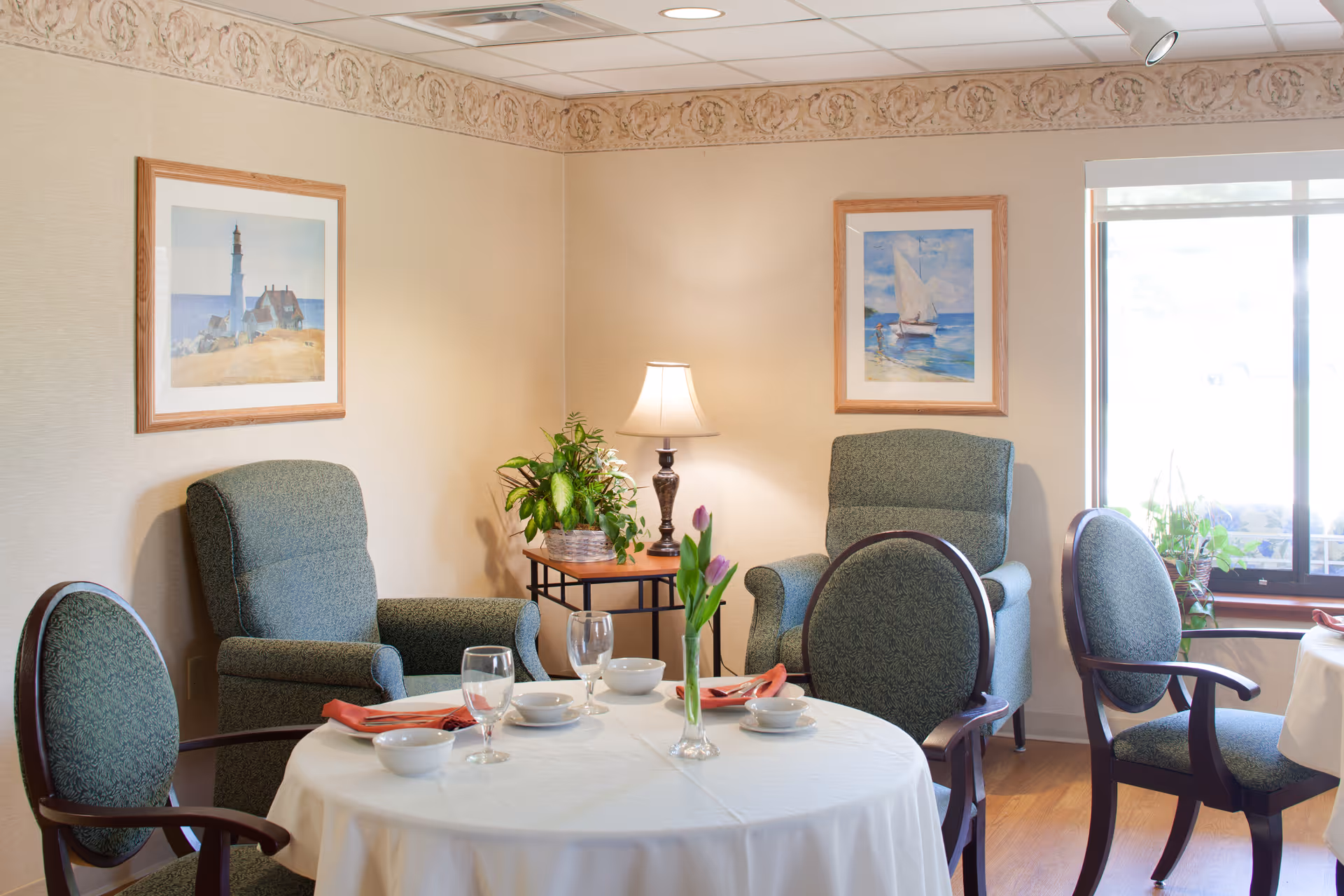 A cozy dining area with a round table covered with a white tablecloth, set with bowls, glasses, and napkins. Four upholstered chairs surround the table. In the background, there are two comfortable armchairs, a side table with a lamp and a potted plant, and framed paintings on the wall. A window with a roller shade lets in natural light.
