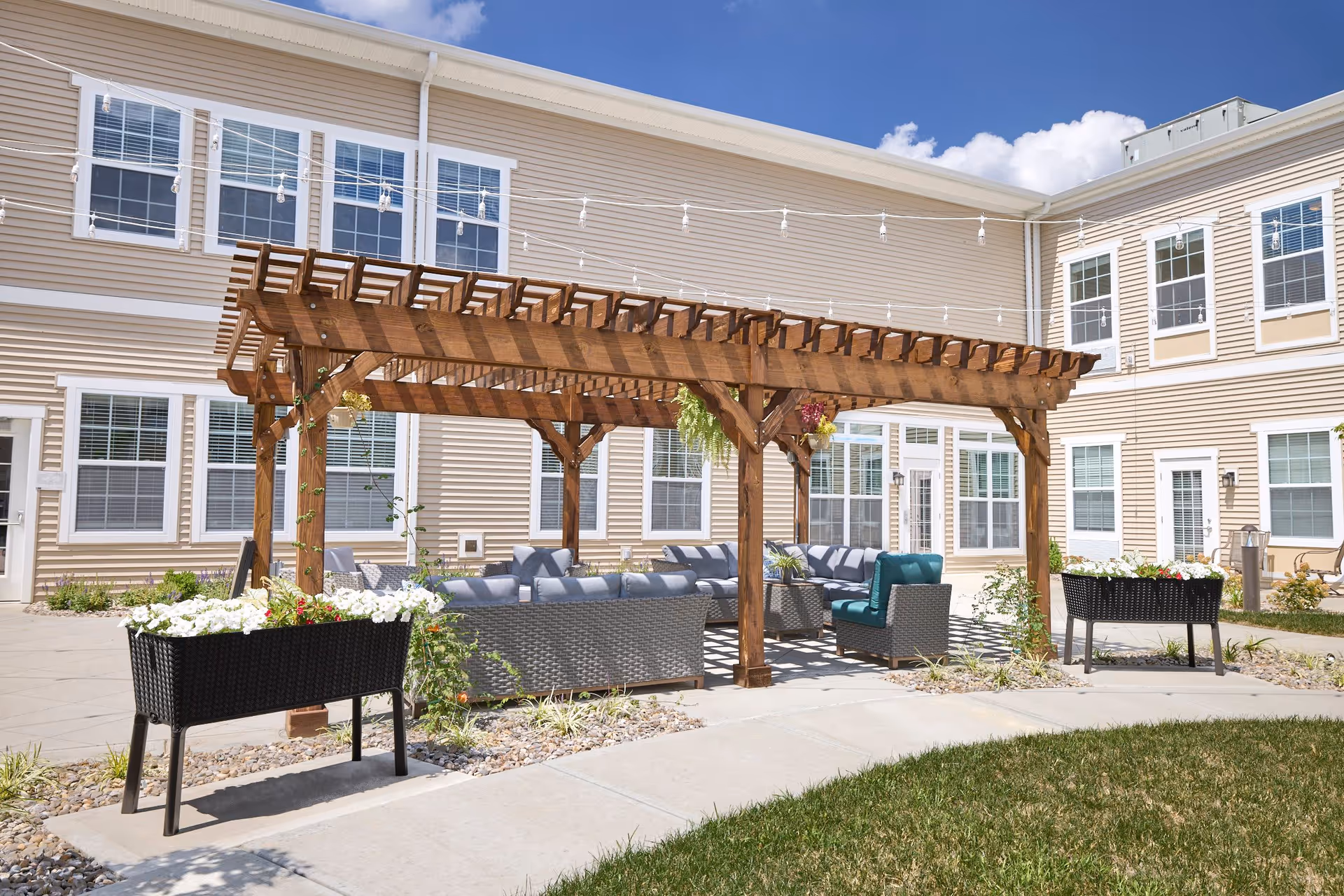 Outdoor seating area at a senior living facility with a wooden pergola, cushioned wicker chairs and sofas arranged underneath, surrounded by planters with white flowers, and a beige building with multiple windows in the background under a clear blue sky.
