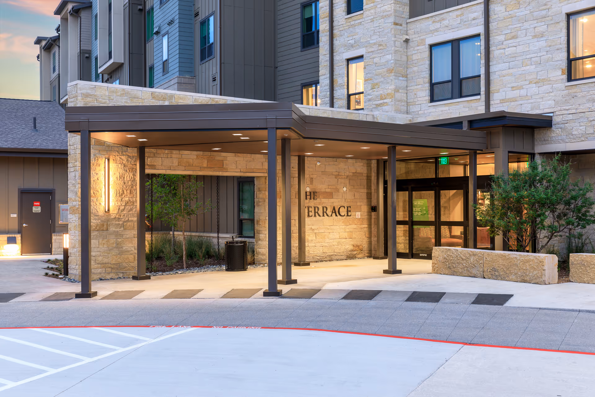 Entrance area of a senior living facility named The Terrace at Touchmark at Emerald Lake, featuring a covered drop-off zone with stone walls, modern lighting, glass doors, and surrounding landscaping with bushes and small trees.