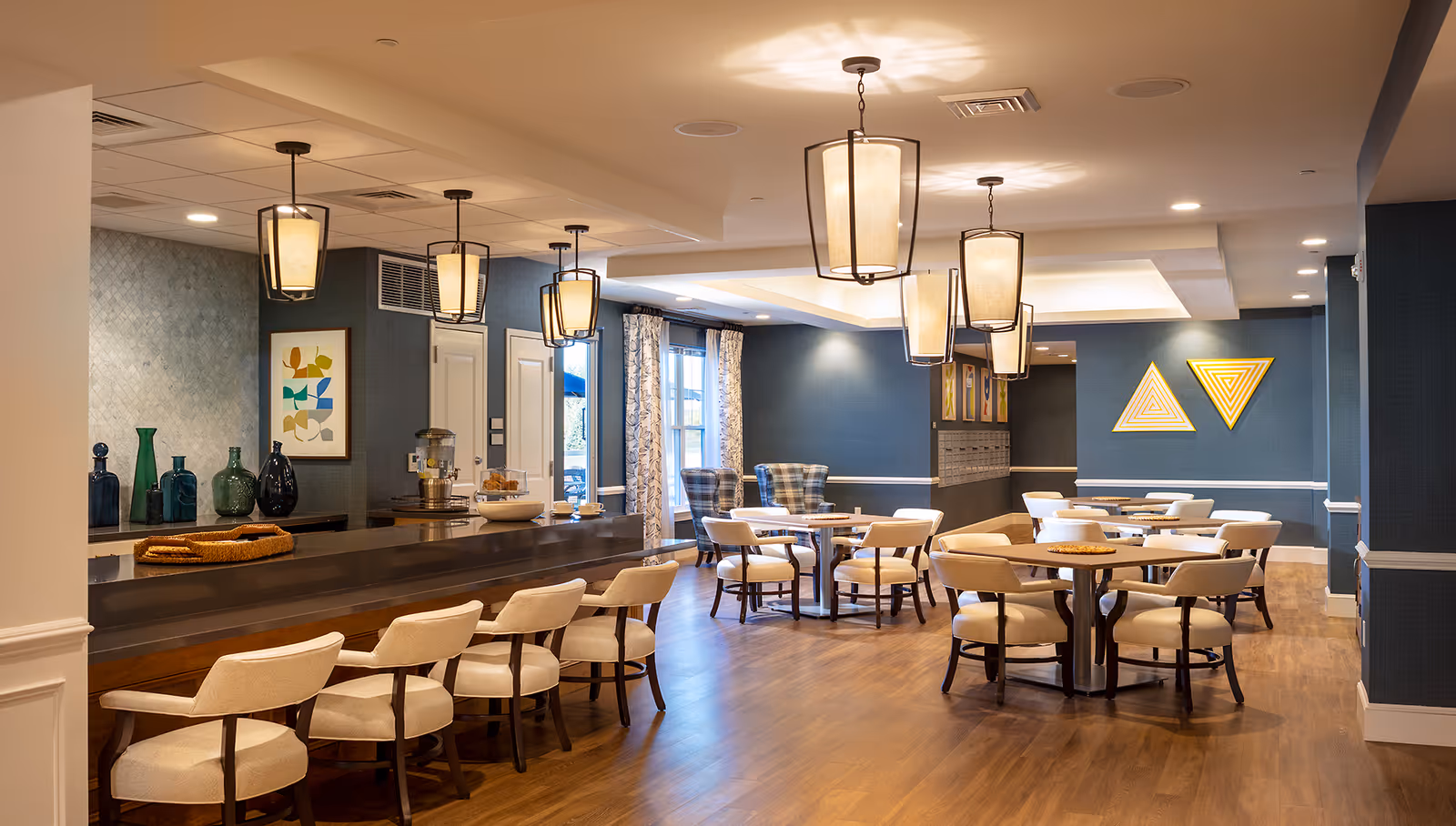 A well-lit dining area in a senior living facility featuring multiple round tables with beige cushioned chairs, a long counter with high chairs, modern pendant lights hanging from the ceiling, blue textured walls with framed abstract art, and large windows with patterned curtains allowing natural light to enter.