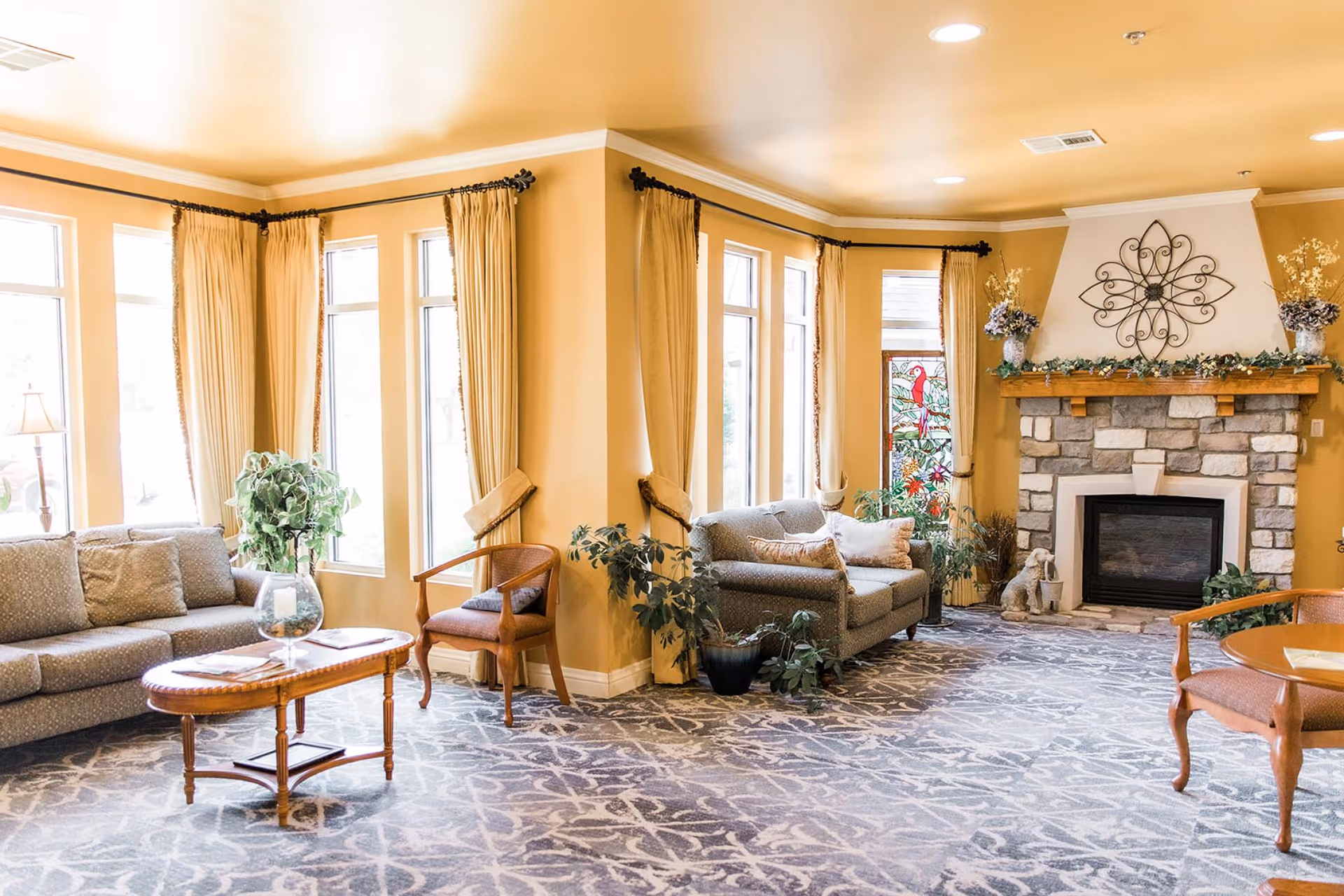 Sunny communal living room with sofas, armchairs, coffee tables, potted plants, large windows with curtains, and a stone fireplace.