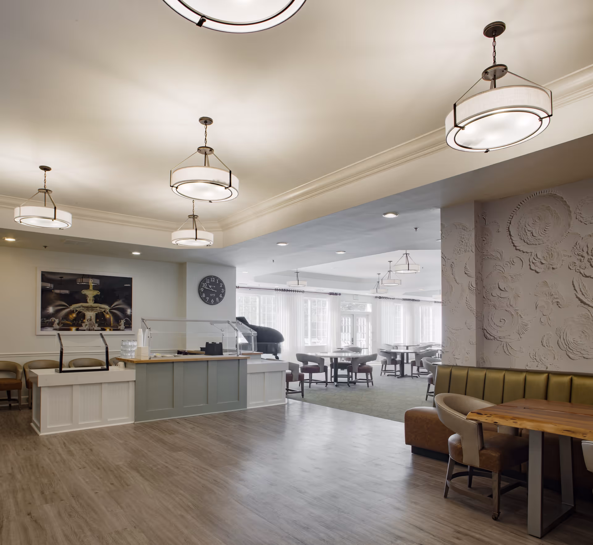 A spacious and well-lit dining area in a senior living facility featuring modern pendant lights, a serving counter with chairs, a wall clock, and multiple tables and chairs arranged near large windows allowing natural light to fill the room. There is also a decorative wall with floral patterns and a wooden table with cushioned chairs in the foreground.