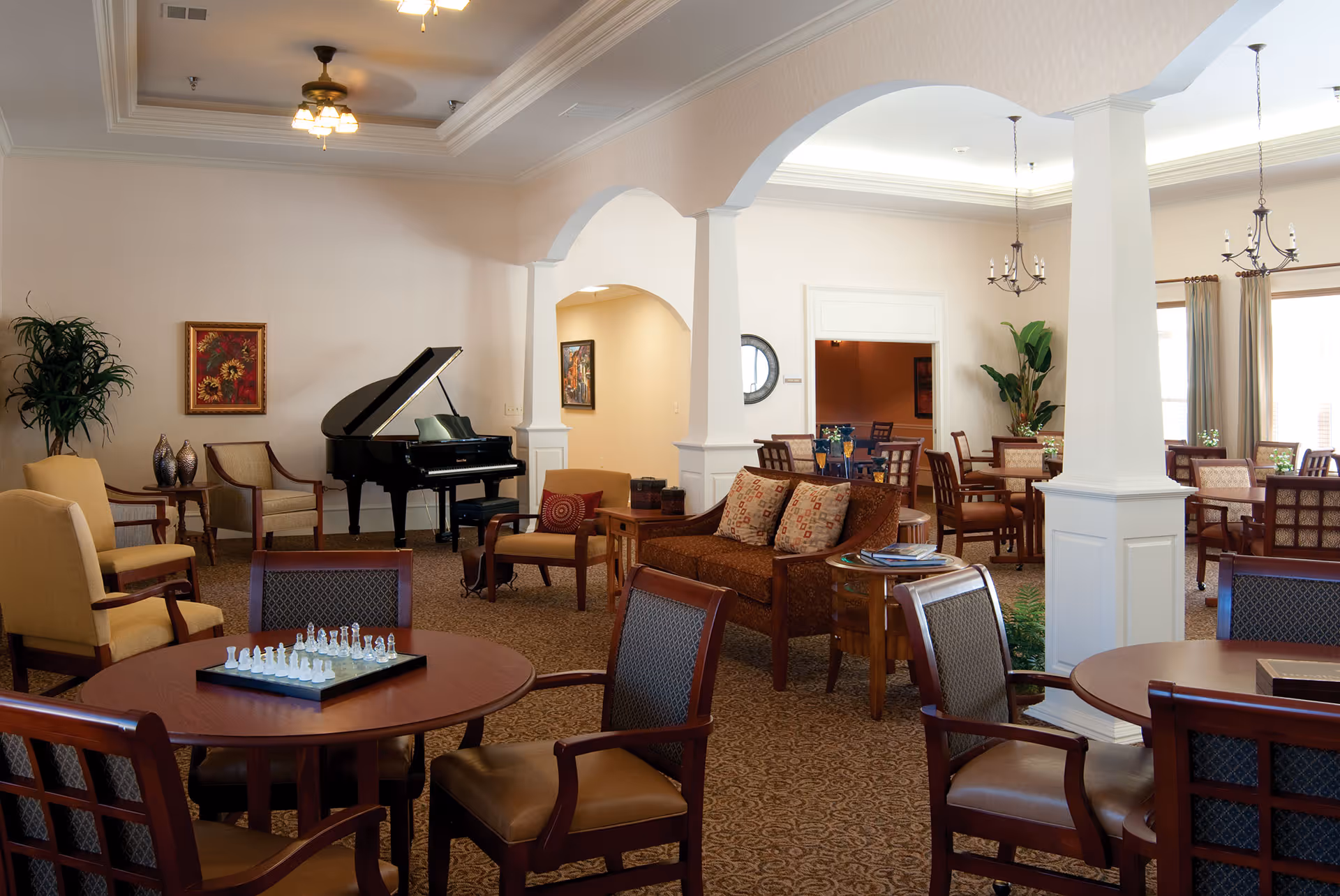 A spacious and well-lit common area in a senior living facility featuring multiple seating arrangements with wooden chairs and tables. A chess set is placed on one of the tables in the foreground. There is a grand piano against the wall, decorative plants, framed artwork, and arched columns dividing the space. The area has carpeted floors and ceiling fans with lights.