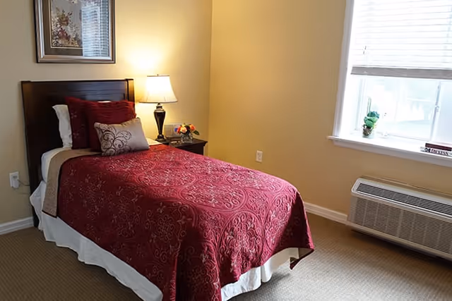 A single bed with a dark wooden headboard, covered with a red patterned bedspread and several pillows, next to a nightstand with a lamp and a small flower arrangement. A window with blinds and a heating/cooling unit is on the right wall.