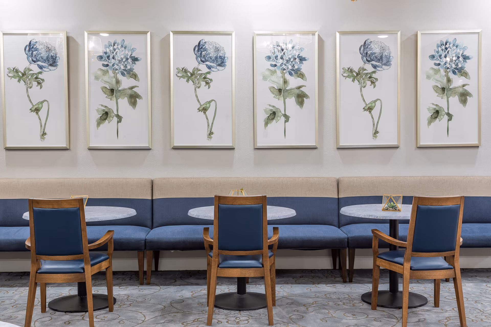 A seating area with three round tables and six wooden chairs with blue cushions arranged in front of a long blue and beige upholstered bench. Above the bench, there are six framed botanical watercolor paintings of blue and green flowers evenly spaced on a white wall.