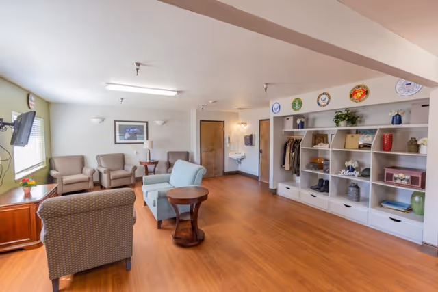 Well-lit communal lounge with armchairs, a sofa, side tables, a wall-mounted TV, and open shelving displaying decor on wood floors.