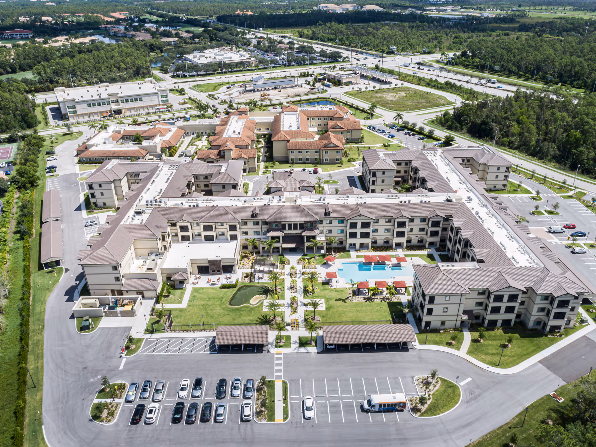 Aerial view of Discovery Village At Naples senior living facility showing multiple connected buildings with beige and brown roofs surrounding a central courtyard that includes a swimming pool, lounge chairs, shaded seating areas, and landscaped green spaces. There are parking lots with cars and a shuttle bus in front of the facility, and surrounding roads and greenery are visible in the background.
