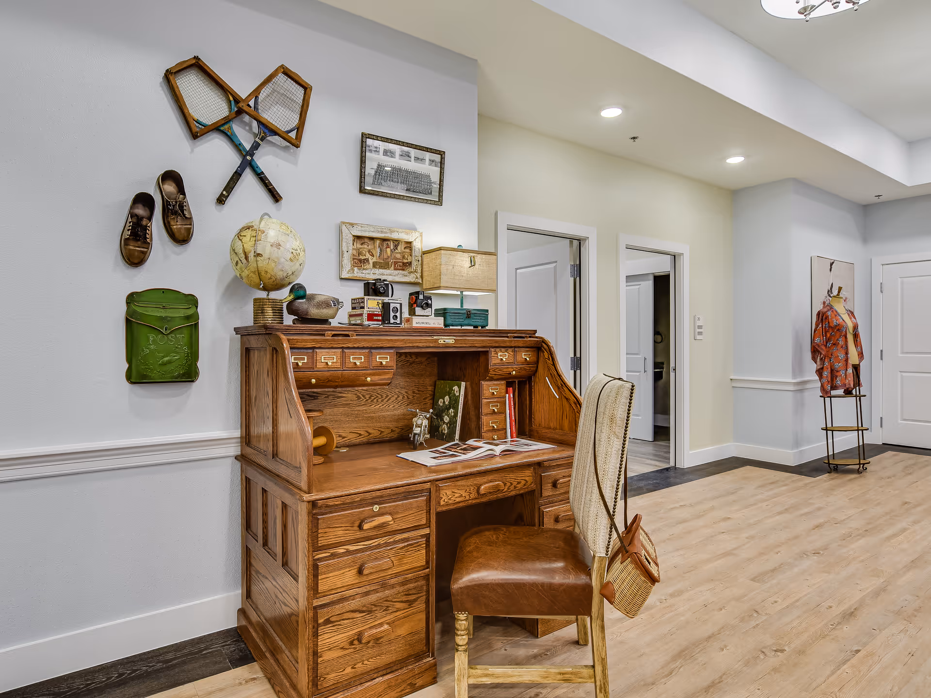 A wooden roll-top desk with a leather chair and decorative wall accents in a bright senior living common area.