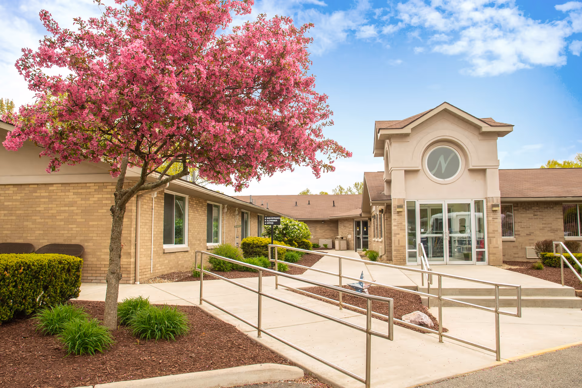 Front entrance of a brick senior living facility with a pink flowering tree, paved walkways and accessible ramps leading to glass doors.