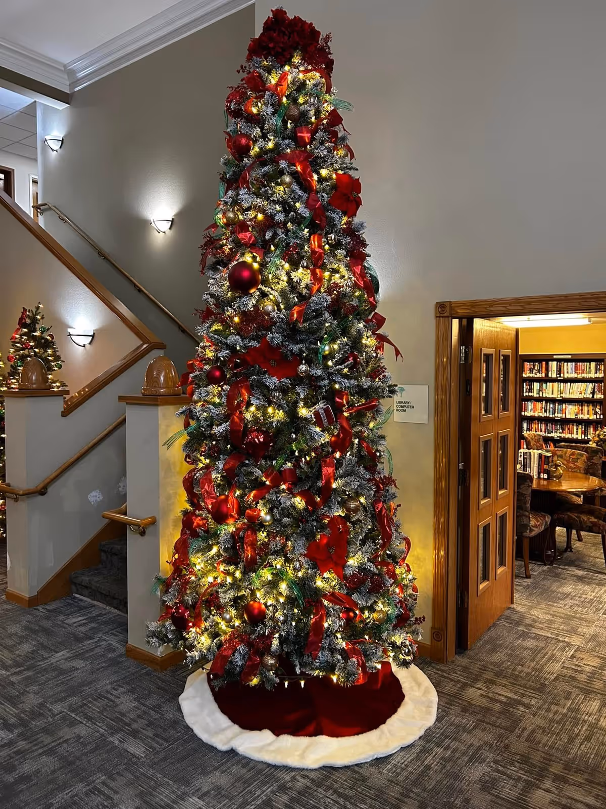 A tall, decorated Christmas tree with red ribbons, ornaments, and lights stands in a carpeted interior space near a staircase and an open doorway leading to a room filled with bookshelves and chairs.
