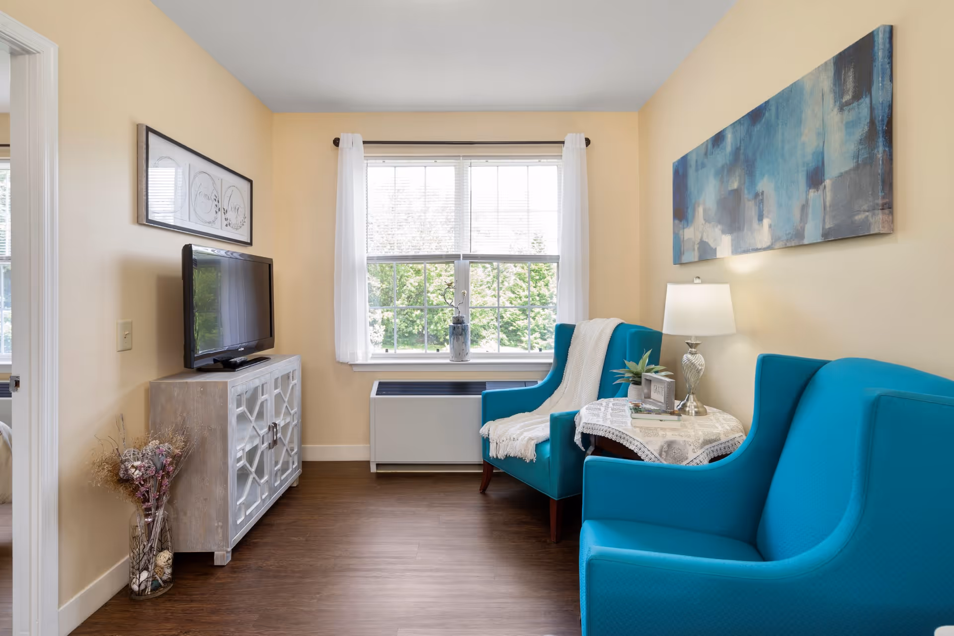 A cozy living room with two bright blue armchairs, a small round table with a lace tablecloth, a lamp, and decorative items. A large window with white curtains lets in natural light, and a TV sits on a cabinet against the wall. The walls are painted light yellow, and there is a blue abstract painting hanging above one of the chairs.