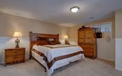 A cozy bedroom with a large wooden bed featuring white and brown bedding. There are two wooden nightstands on either side of the bed, each with a lamp. A wooden dresser and armoire are positioned against the wall. The room has beige walls and carpeted flooring, with soft lighting creating a warm atmosphere.