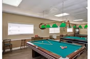 Recreation room interior featuring two green-felt pool tables with hanging green billiard lights, windows with blinds, and seating.