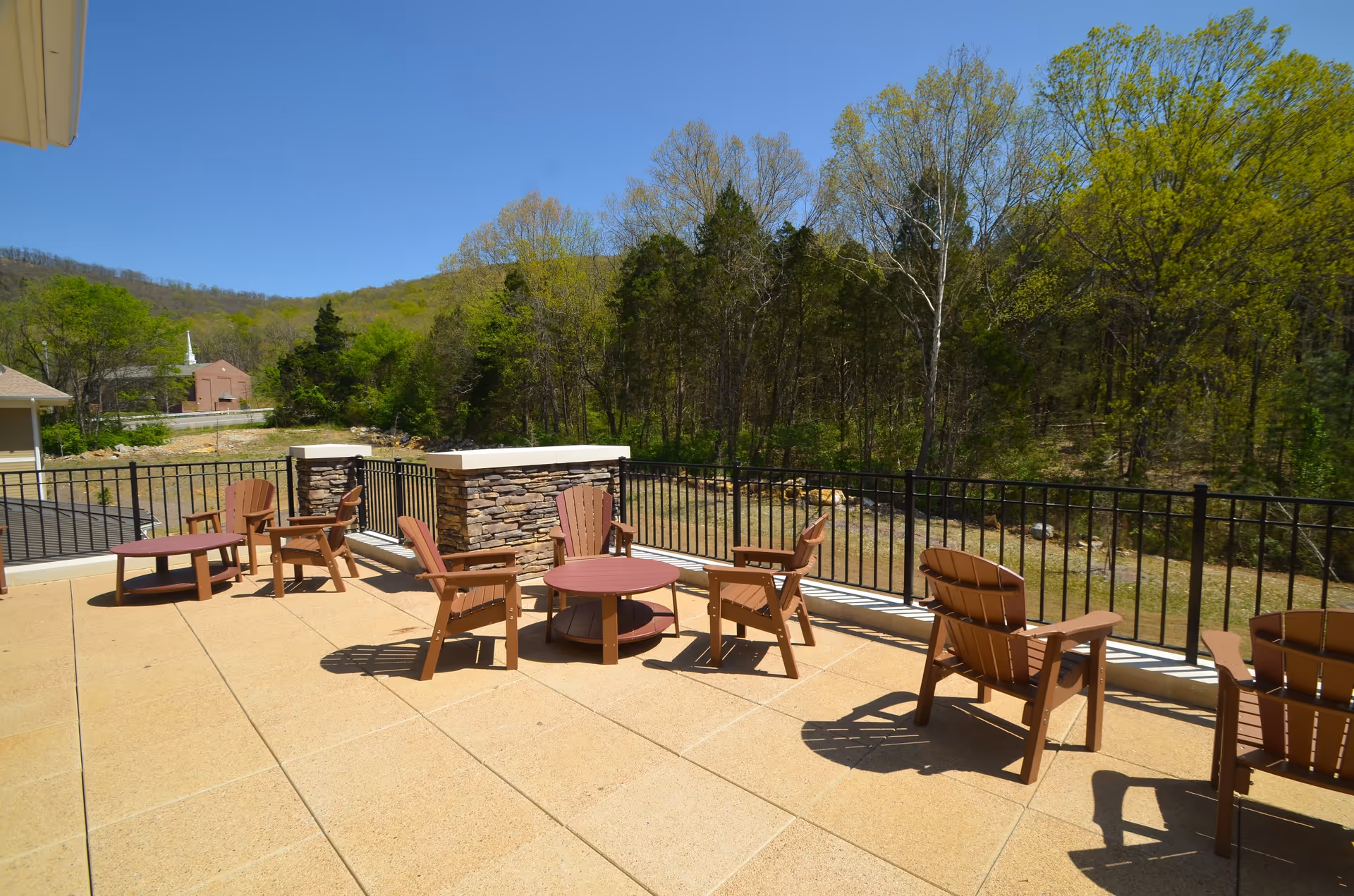 Outdoor patio area with several wooden chairs and round tables on a tiled floor, surrounded by a black metal railing. In the background, there are trees and a clear blue sky.