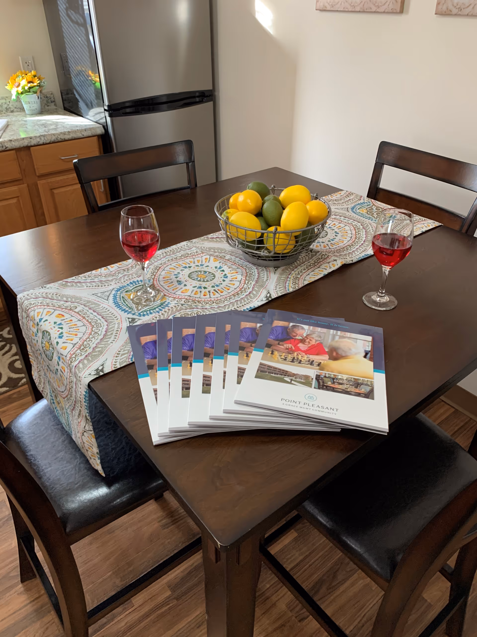 A wooden dining table with a colorful patterned runner, a wire basket filled with lemons and limes, two glasses of red beverage, and several brochures for Point Pleasant arranged on the table. The background shows part of a kitchen with wooden cabinets and a stainless steel refrigerator.