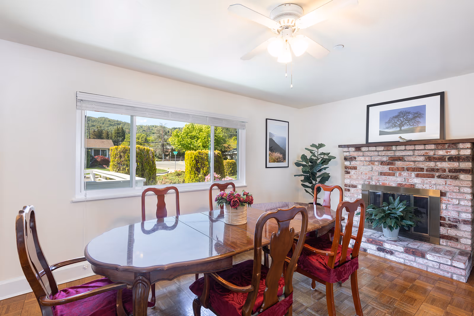 A bright dining room with a wooden dining table and six chairs with red cushions. A flower arrangement is placed in the center of the table. There is a large window showing greenery outside, a ceiling fan with lights, a brick fireplace with a potted plant on the hearth, and framed pictures on the walls.