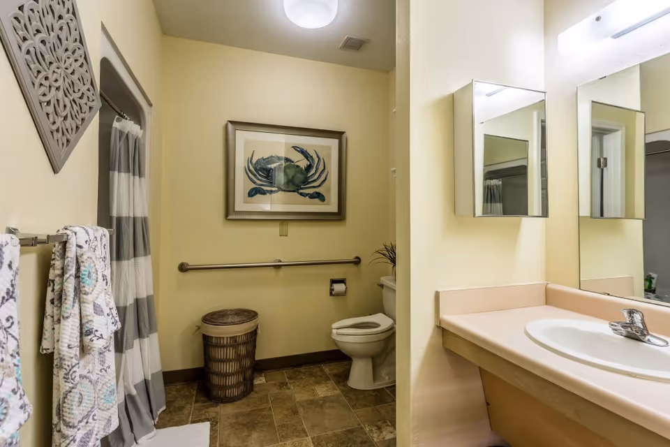 A bathroom with a toilet, a sink with a countertop, a mirror cabinet, a towel rack with patterned towels, a striped shower curtain, a wicker laundry basket, and a framed picture of a blue crab on the wall.