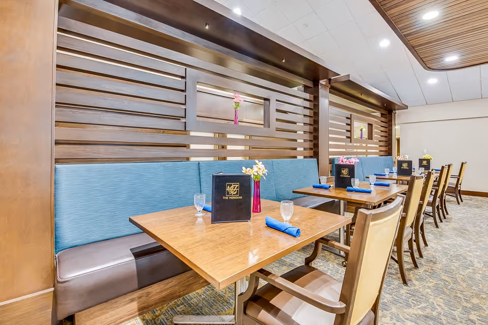 Interior view of a dining area in a senior living facility with wooden tables, blue cushioned bench seating, and wooden chairs. Each table has a menu, a glass of water, and a blue napkin. The background features wooden panel dividers with small flower vases placed in openings. The ceiling has recessed lighting and a wooden accent panel.
