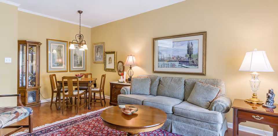 A cozy living room and dining area with a gray sofa, wooden coffee table, and a wooden dining table with four chairs. The walls are painted light yellow and decorated with framed artwork. There are two table lamps on wooden side tables and a hanging light fixture above the dining table. A patterned rug covers part of the wooden floor.