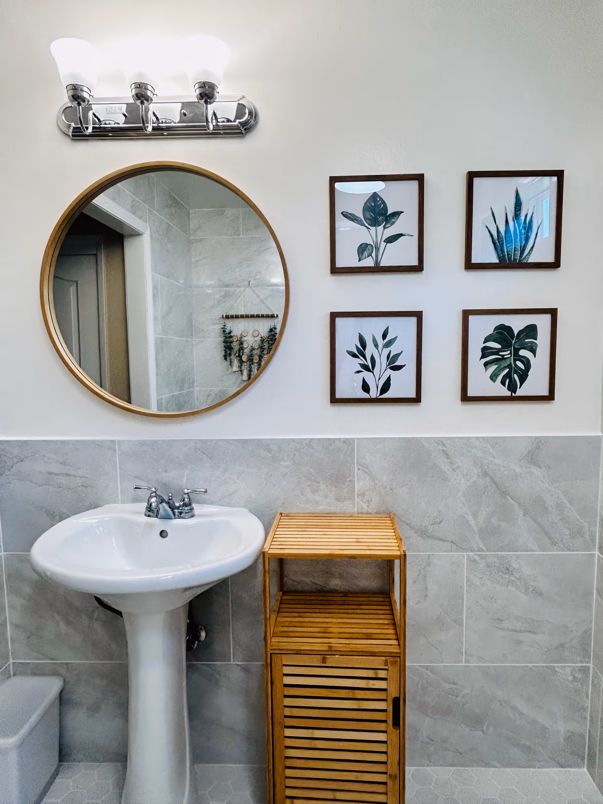 Small modern bathroom with a pedestal sink, round mirror, wall light, four framed plant prints and a wooden storage cabinet.