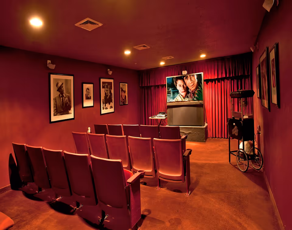 A small theater room with two rows of red theater seats facing a television screen showing a movie scene. The walls are painted red and decorated with framed black and white portraits. A vintage popcorn machine is positioned on the right side of the room.