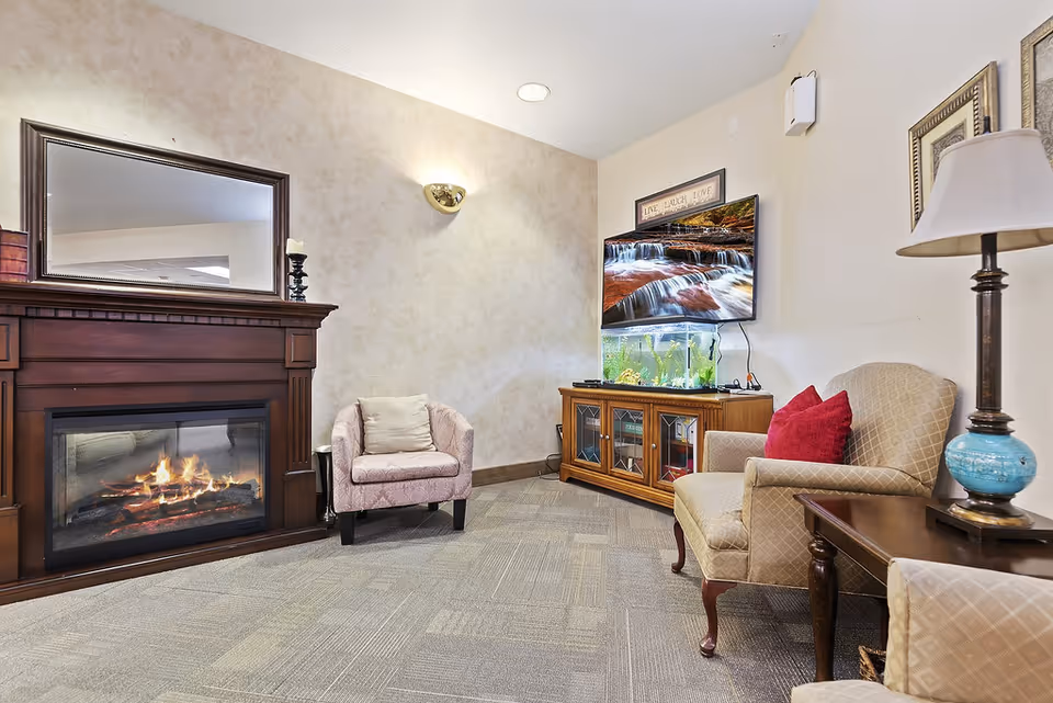 A cozy living room area featuring a lit fireplace with a wooden mantel and a large mirror above it. There are two upholstered armchairs, one with a red pillow, a wooden side table with a lamp, and a wooden cabinet with a fish tank and a wall-mounted TV displaying a waterfall scene. The walls are light-colored, and the floor is carpeted.