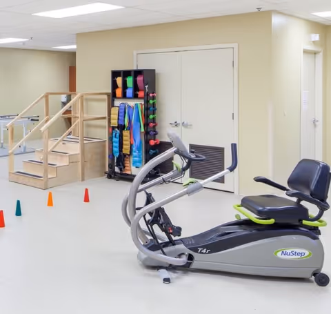A rehabilitation room featuring a NuStep T4r recumbent cross trainer exercise machine, a set of small colorful cones on the floor, a wooden staircase with handrails, and a black rack holding various exercise bands and weights against a beige wall with double doors.