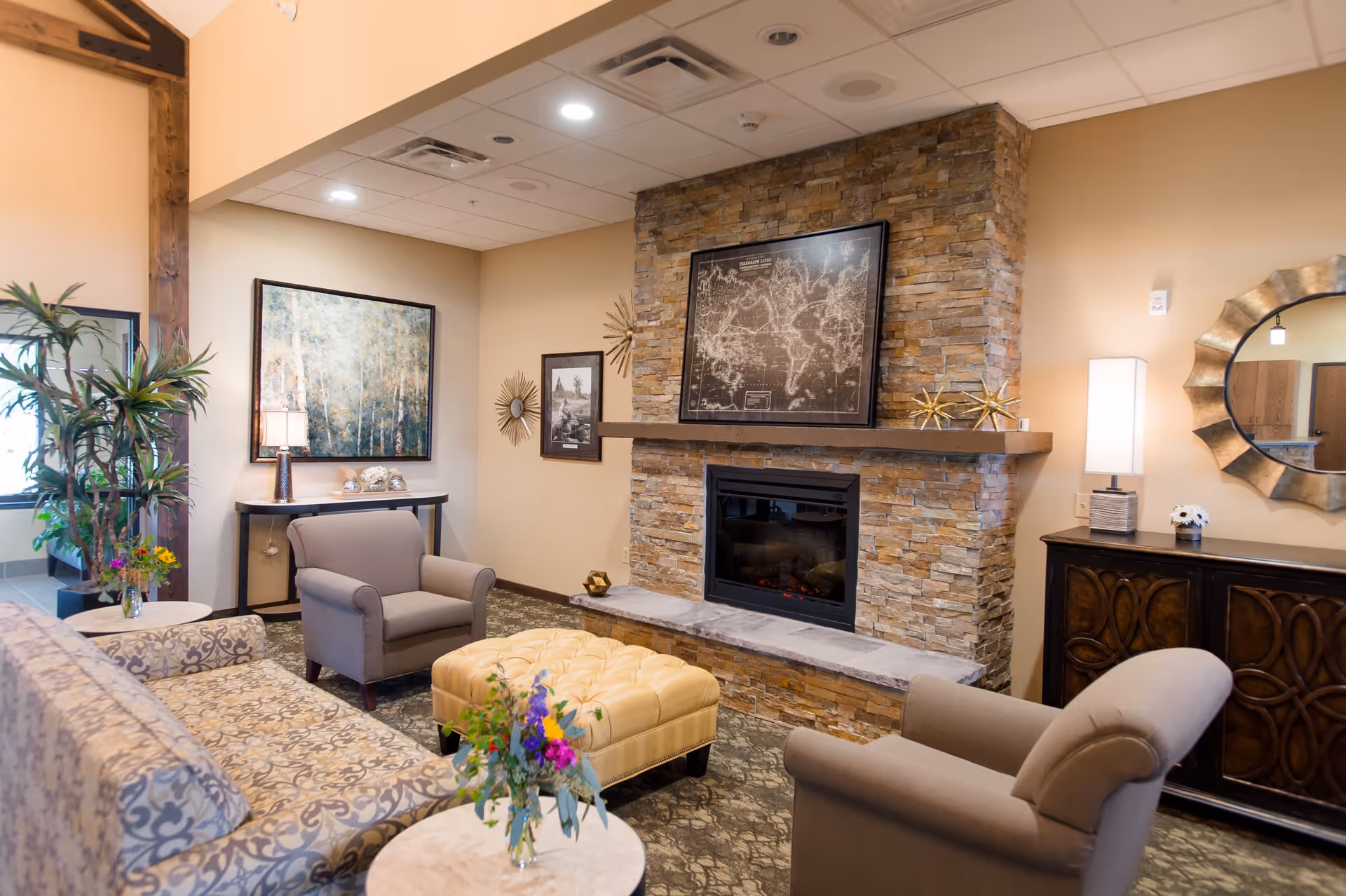 A cozy senior living community lounge area with a stone fireplace, a framed map above the mantel, two beige armchairs, a patterned sofa, a tufted ottoman, side tables with flower arrangements, a console table with a lamp and decorative items, and a large round mirror on the wall.