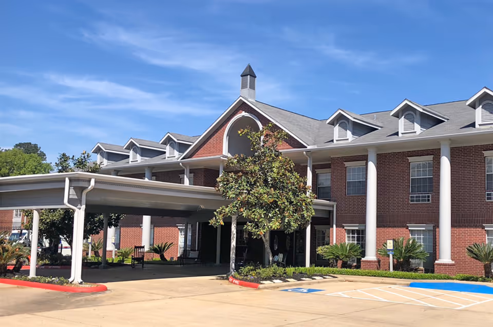 Exterior view of a two-story brick building with white columns and a covered entrance driveway. The building has multiple windows and dormer windows on the roof. There are small landscaped areas with plants and a tree near the entrance. The sky is clear and blue.