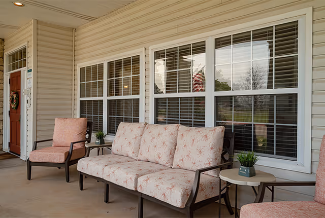 A covered outdoor patio area with cushioned seating including a sofa and two armchairs, small side tables with potted plants, and large windows with white blinds on the building wall behind.