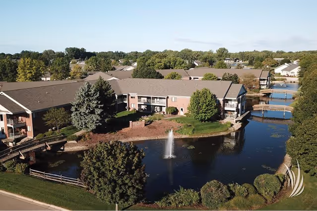 Aerial view of Royal Park Place Retirement Living showing a large residential building complex surrounding a pond with a water fountain. The area features green trees, shrubs, and wooden bridges crossing the water. The sky is clear with some clouds.