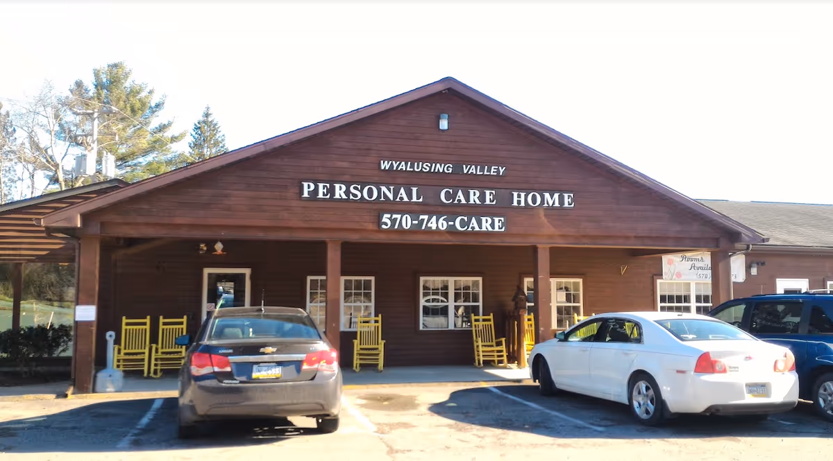 Front exterior view of Wyalusing Valley Personal Care Home building with a wooden facade, a covered porch with yellow rocking chairs, and three cars parked in front.