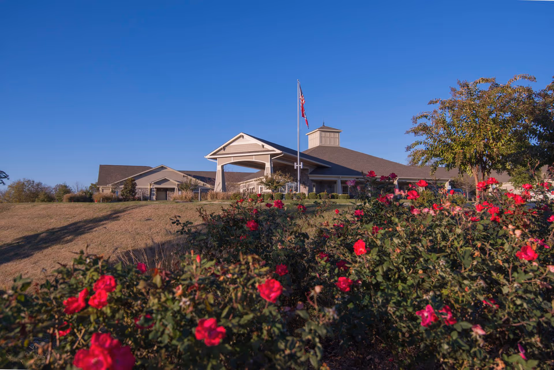 Exterior view of Morning Pointe of Brentwood Assisted Living facility with a clear blue sky, a flagpole with American and state flags, a landscaped garden with blooming flowers, and a tree on the right side.