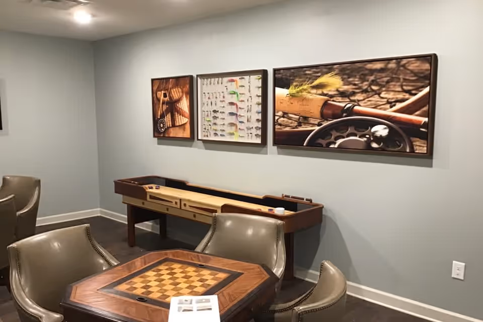 A cozy game room with a wooden chess/checkers table surrounded by four leather chairs. On the wall behind, there is a shuffleboard table and three framed pictures featuring fishing-themed images.