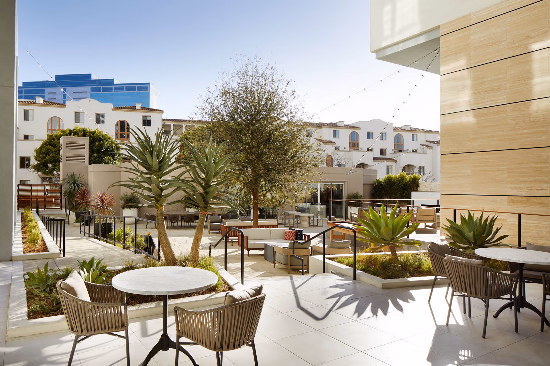Outdoor courtyard patio with tables, chairs, planters, trees and string lights between buildings.