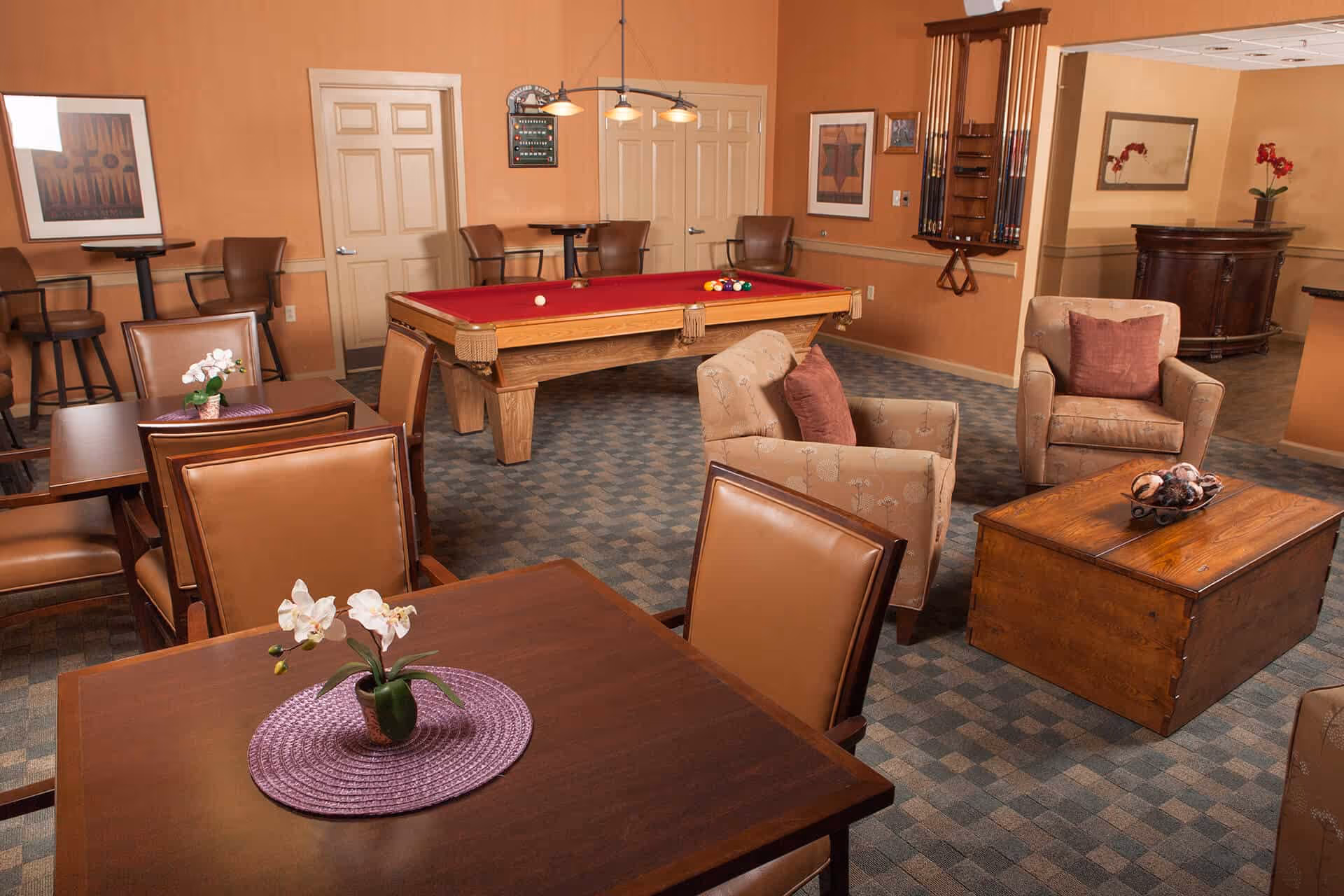 A cozy common room with a red felt pool table in the center, surrounded by several brown leather chairs and tables with small flower arrangements. There are two beige armchairs with red cushions facing a wooden coffee table. The walls are painted a warm orange color and decorated with framed artwork and a rack holding pool cues. The room has a carpeted floor with a checkered pattern.