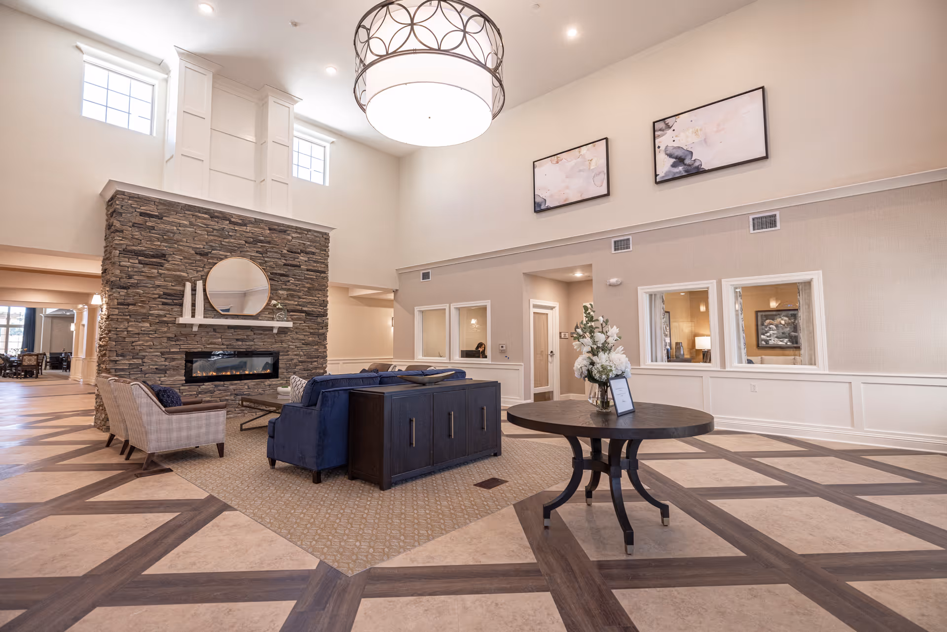 Spacious senior living facility common area with high ceilings, a large stone fireplace, comfortable seating including a blue sofa and two armchairs, a round table with a floral arrangement, and modern artwork on the walls.