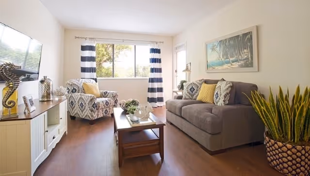 A bright living room with a large window covered by white curtains with navy blue stripes. The room features a gray sofa with patterned and yellow throw pillows, a patterned armchair with a yellow pillow, a wooden coffee table with decorative items, a white TV stand with a mounted flat-screen TV, and a large potted plant. A framed beach-themed painting hangs on the wall above the sofa.