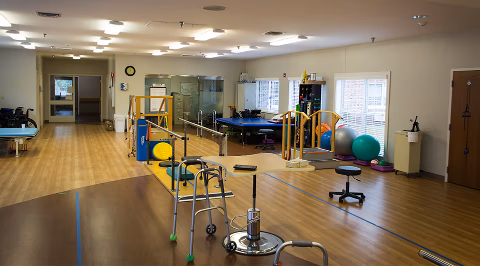 Spacious rehabilitation/therapy room with parallel bars, walkers, exercise balls, treatment tables, and stools on wood flooring.