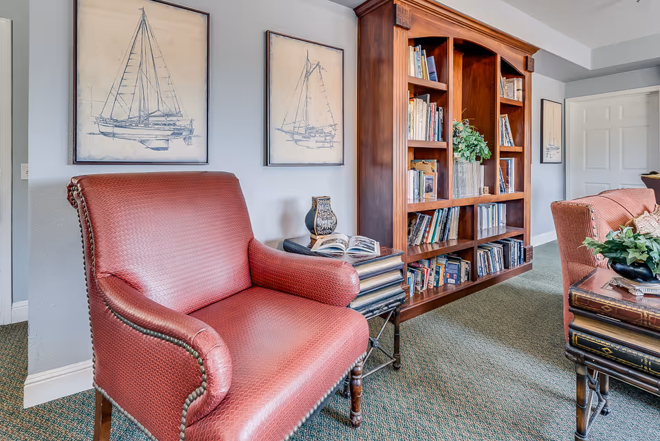 A cozy living room area featuring a red upholstered armchair, a wooden bookshelf filled with books and decorative items, and framed nautical-themed artwork on the light gray walls. The room has a green carpet and a white door in the background.