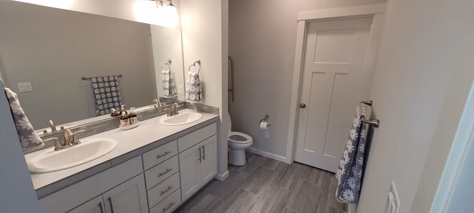 A clean, modern bathroom with a double sink vanity featuring white cabinets and silver faucets. Above the sinks is a large mirror with two light fixtures. The bathroom has gray tiled flooring, a white toilet, and several gray towels with white floral patterns hanging on towel racks.
