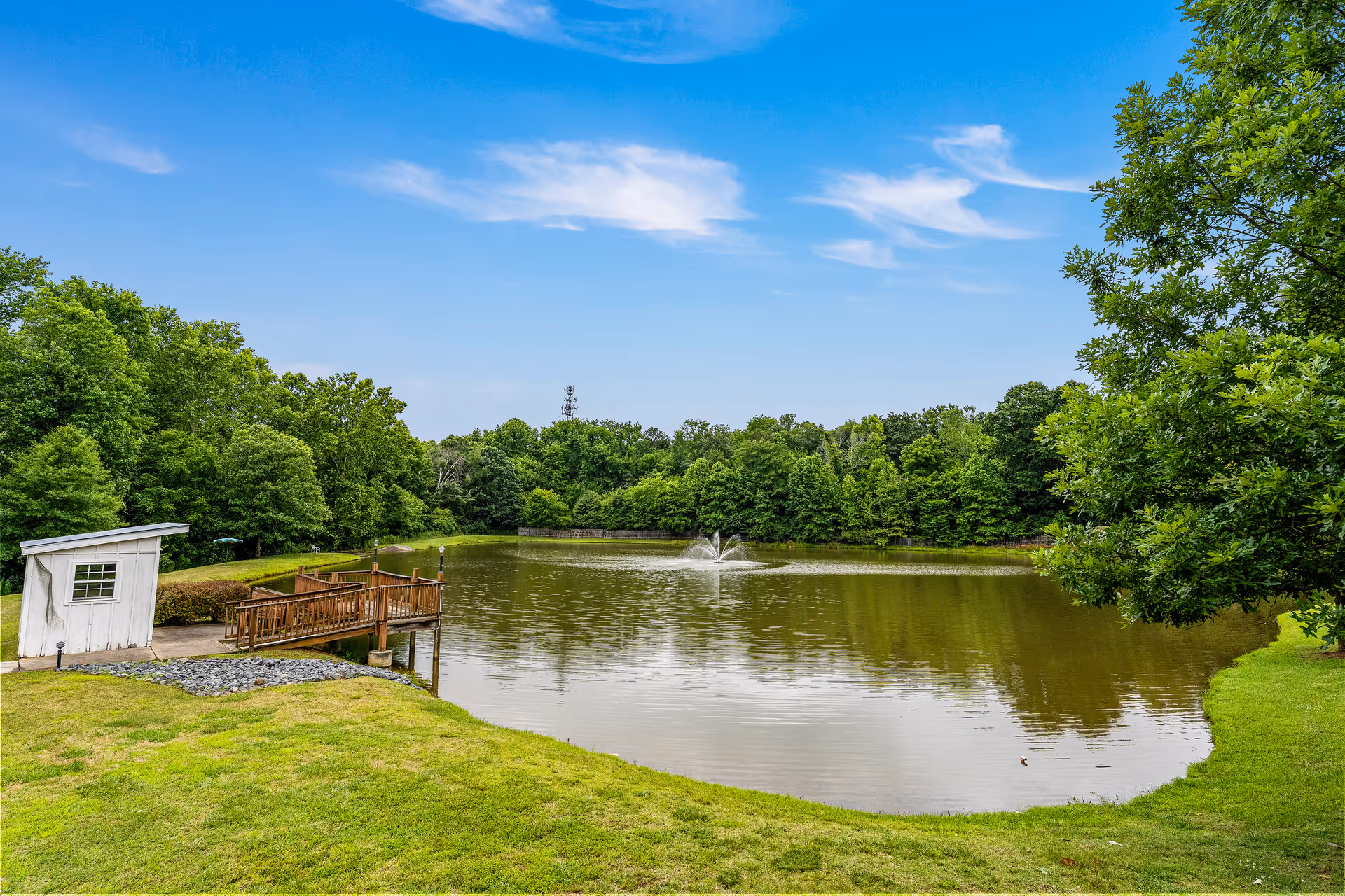 A serene outdoor scene featuring a small pond with a water fountain in the center, surrounded by lush green trees and grass. There is a wooden dock with railings extending into the pond, and a small white shed near the dock. The sky is clear with a few wispy clouds.
