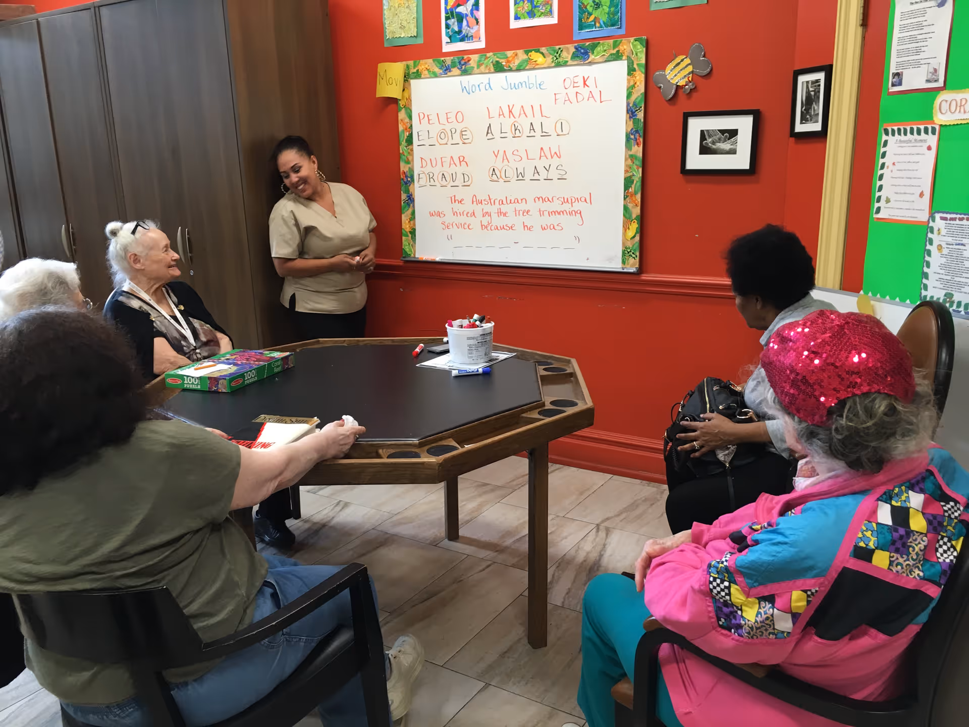 A group of elderly people sitting around a hexagonal table in a room with red walls, engaging in a word jumble activity led by a standing staff member. The whiteboard on the wall displays scrambled words and a sentence to complete. The room has colorful artwork and informational posters on the walls.